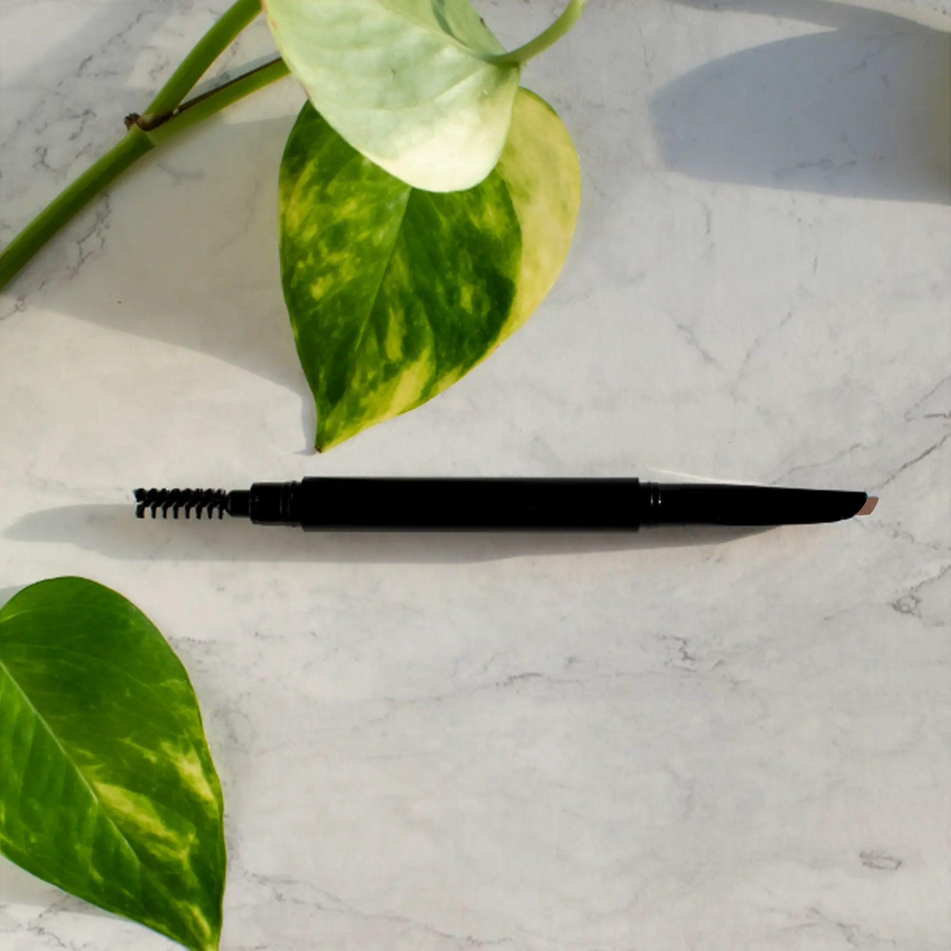 Automatic eyebrow pencil with dual-tip design on a marble surface, surrounded by green leaves.