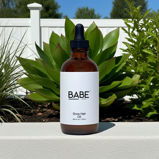 Bottle of Babe Grow Hair Oil with dropper, placed outdoors on a white surface with green plants and white fence background.